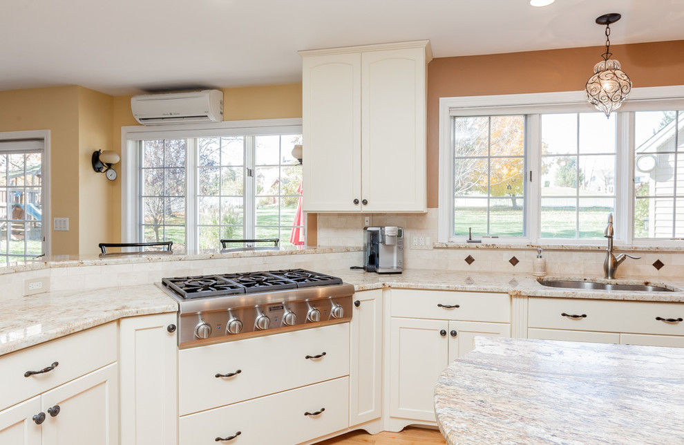 Example of a mid-sized transitional u-shaped light wood floor enclosed kitchen design in Boston with a double-bowl sink, recessed-panel cabinets, white cabinets, granite countertops, beige backsplash, stone tile backsplash, stainless steel appliances and an island