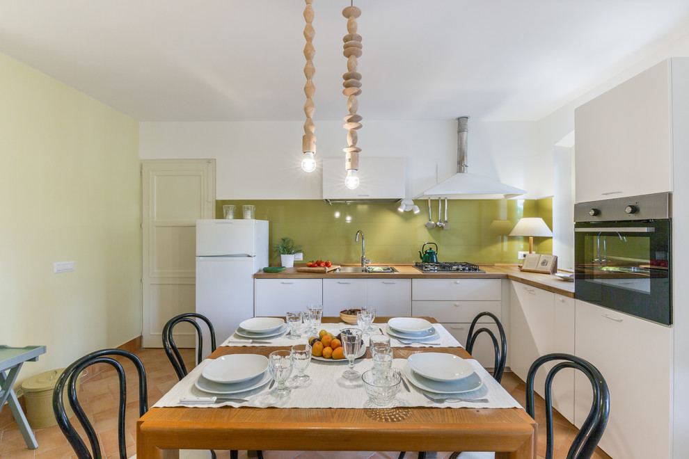 Inspiration for a large country l-shaped eat-in kitchen in Catania-Palermo with a drop-in sink, flat-panel cabinets, white cabinets, wood benchtops, green splashback, ceramic splashback, stainless steel appliances and terra-cotta floors.