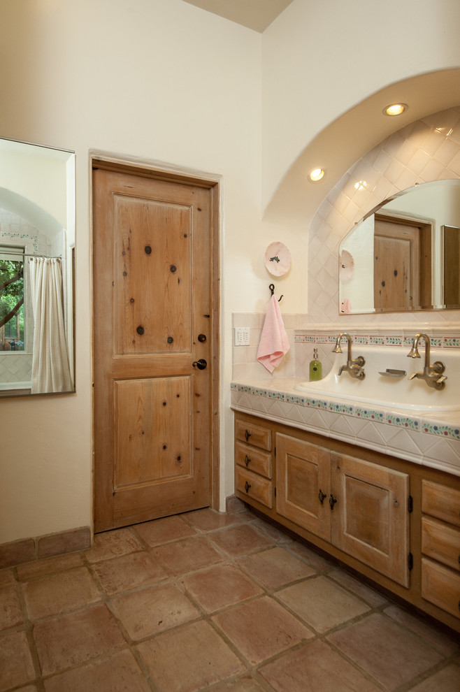 This is an example of a mediterranean bathroom in Other with a trough sink, recessed-panel cabinets, medium wood cabinets, tile benchtops, beige tile, beige walls, terra-cotta floors and beige benchtops.