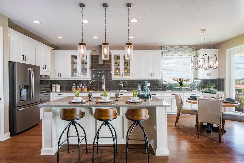 Photo of a country eat-in kitchen in Tampa with a drop-in sink, recessed-panel cabinets, white cabinets, quartzite benchtops, grey splashback, glass tile splashback, stainless steel appliances, medium hardwood floors, with island, brown floor and grey benchtop.