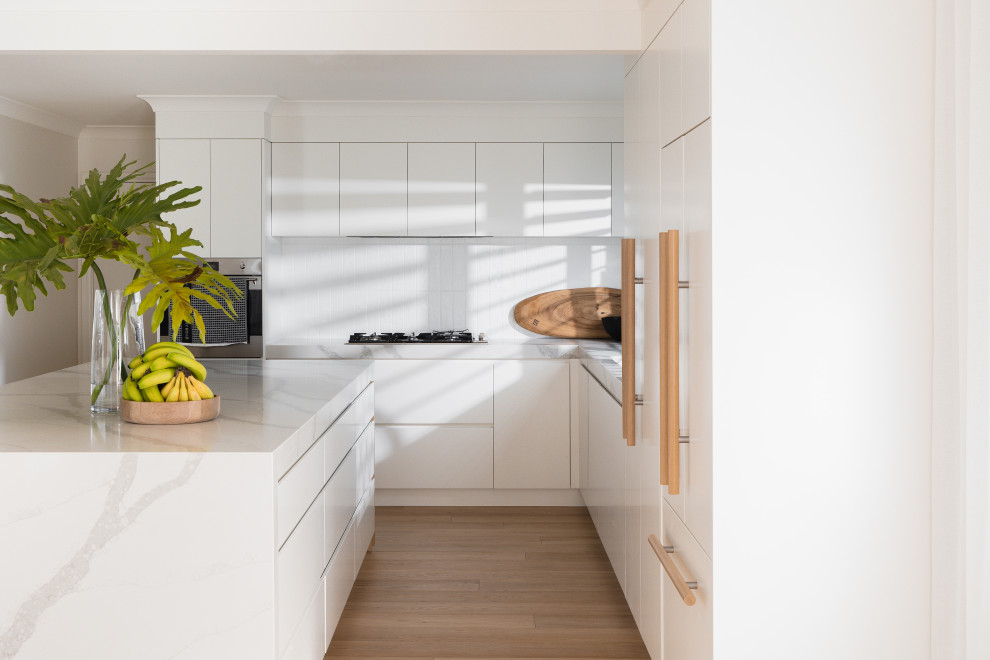 Inspiration for a contemporary l-shaped kitchen in Gold Coast - Tweed with an undermount sink, flat-panel cabinets, white cabinets, white splashback, stainless steel appliances, medium hardwood floors, with island, brown floor and white benchtop.