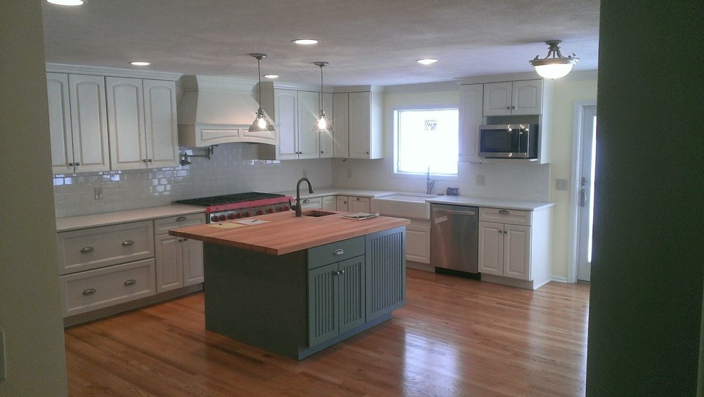 Inspiration for a transitional l-shaped medium tone wood floor kitchen remodel with a farmhouse sink, raised-panel cabinets, white cabinets, white backsplash, subway tile backsplash, stainless steel appliances and an island
