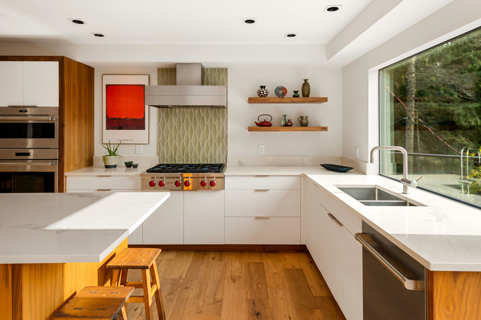 This is an example of a mid-sized midcentury u-shaped eat-in kitchen in Vancouver with an undermount sink, flat-panel cabinets, white cabinets, quartz benchtops, green splashback, glass tile splashback, stainless steel appliances, light hardwood floors, with island, beige floor and white benchtop.