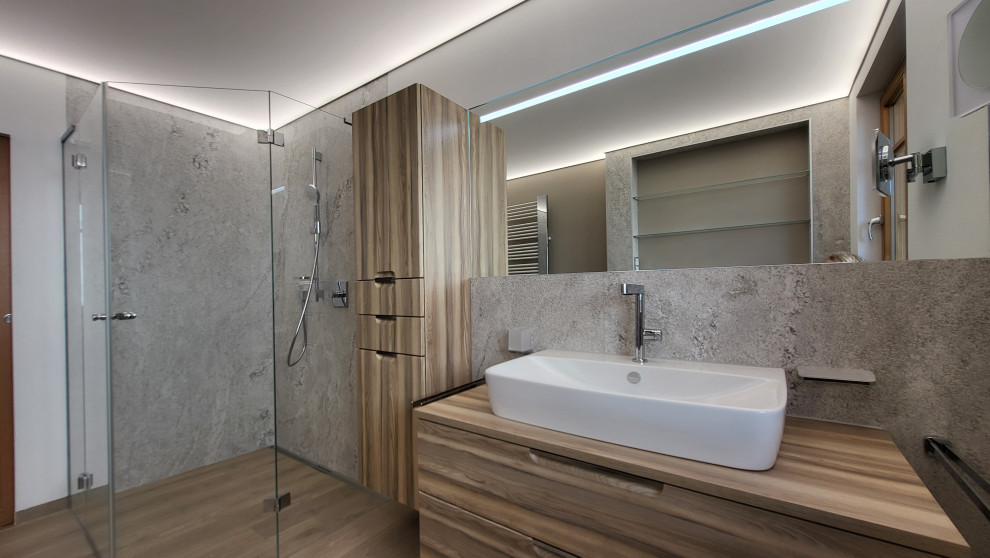 This is an example of a mid-sized country bathroom in Frankfurt with flat-panel cabinets, medium wood cabinets, a curbless shower, a wall-mount toilet, gray tile, stone tile, white walls, wood-look tile, a vessel sink, wood benchtops, brown floor, brown benchtops, a single vanity, a built-in vanity and recessed.