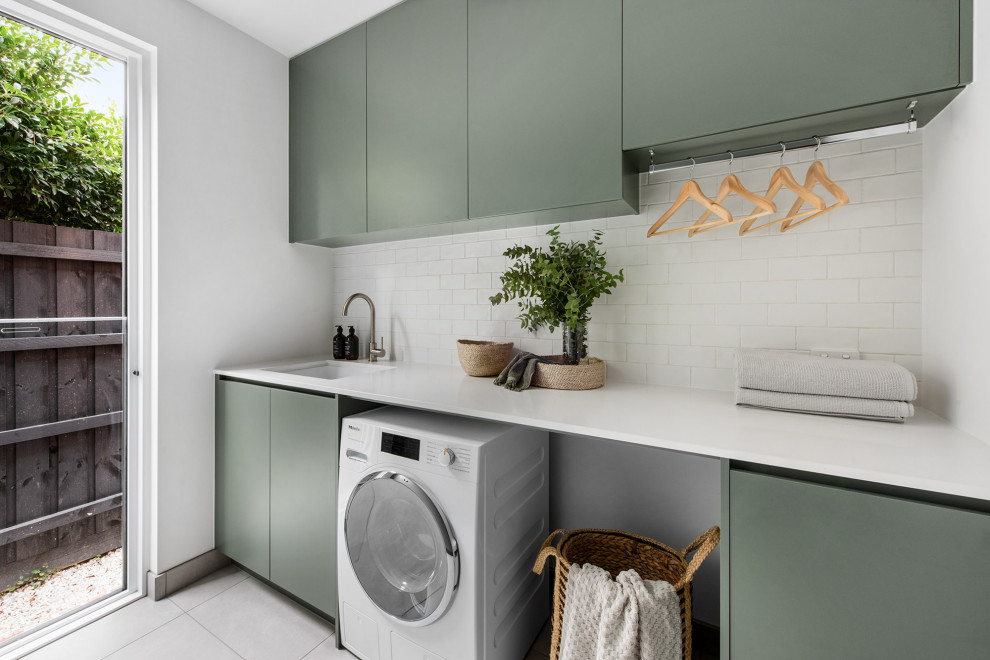 Mid-sized trendy galley porcelain tile and gray floor dedicated laundry room photo in Melbourne with a drop-in sink, flat-panel cabinets, green cabinets, limestone countertops, white backsplash, subway tile backsplash, white walls, an integrated washer/dryer and white countertops