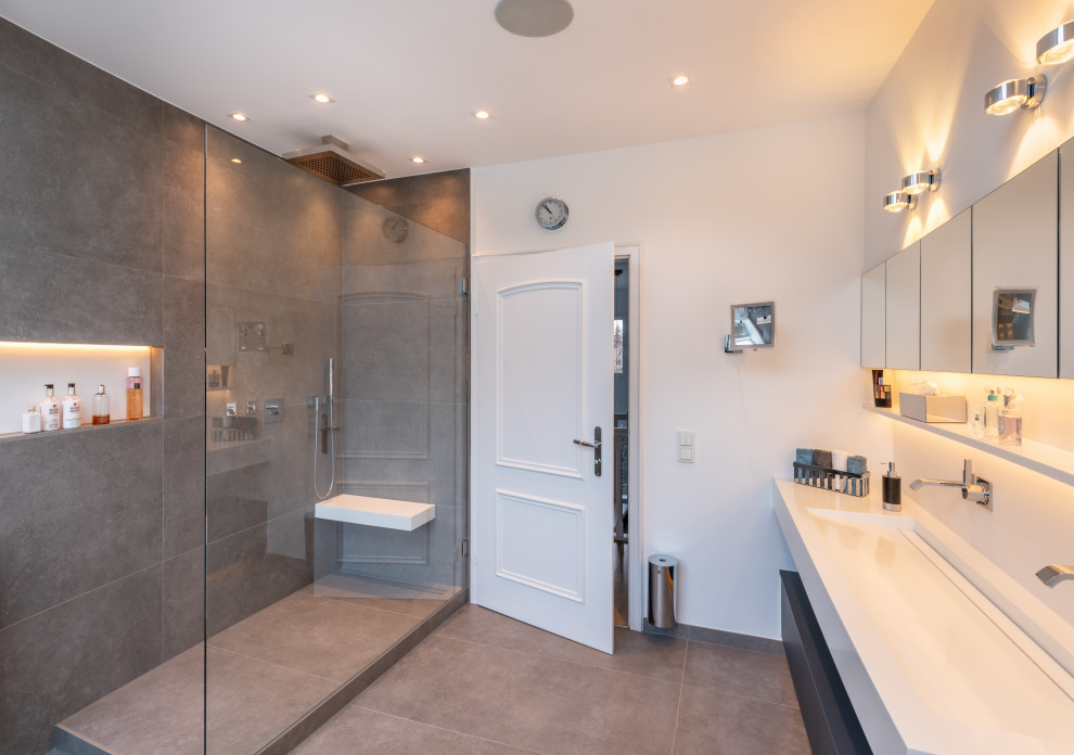 Contemporary bathroom in Dusseldorf with flat-panel cabinets, grey cabinets, an open shower, gray tile, ceramic tile, white walls, ceramic floors, a wall-mount sink, solid surface benchtops, grey floor, an open shower, white benchtops, a shower seat, a double vanity and a floating vanity.