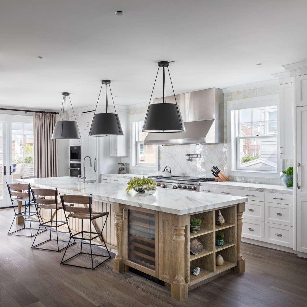 Inspiration for a beach style galley open plan kitchen in New York with a farmhouse sink, recessed-panel cabinets, white cabinets, marble benchtops, white splashback, stainless steel appliances, medium hardwood floors, with island and brown floor.