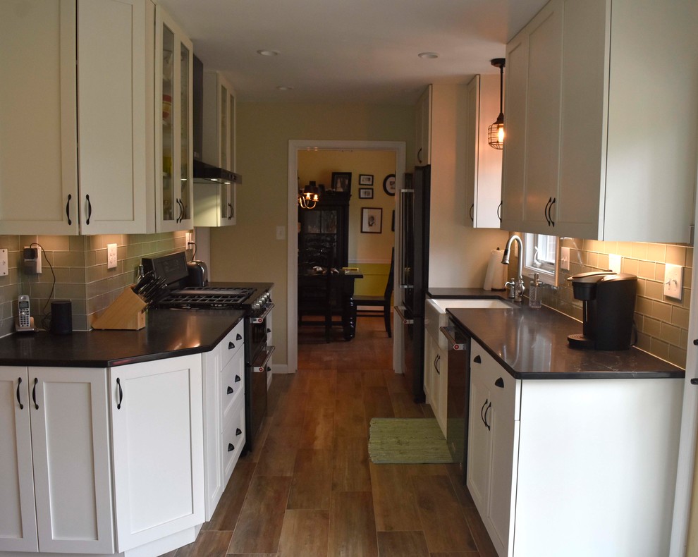 Mid-sized transitional galley laminate floor and brown floor enclosed kitchen photo in DC Metro with a farmhouse sink, shaker cabinets, white cabinets, granite countertops, gray backsplash, subway tile backsplash, stainless steel appliances, no island and black countertops