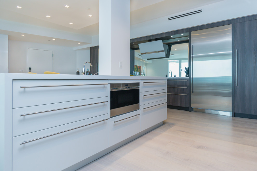 This is an example of a large contemporary l-shaped open plan kitchen in Miami with an undermount sink, shaker cabinets, brown cabinets, solid surface benchtops, mirror splashback, stainless steel appliances, light hardwood floors, with island, beige floor and white benchtop.