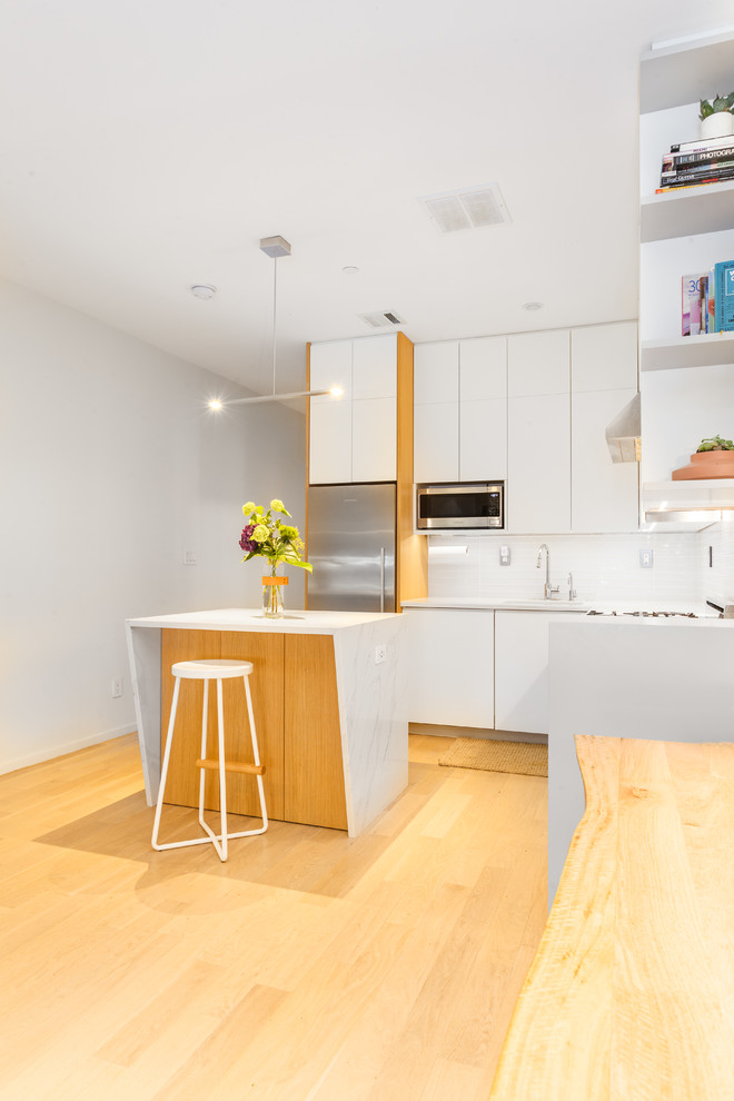 Photo of a small modern l-shaped open plan kitchen in New York with an undermount sink, flat-panel cabinets, white cabinets, quartzite benchtops, white splashback, stainless steel appliances, light hardwood floors, with island and beige floor.