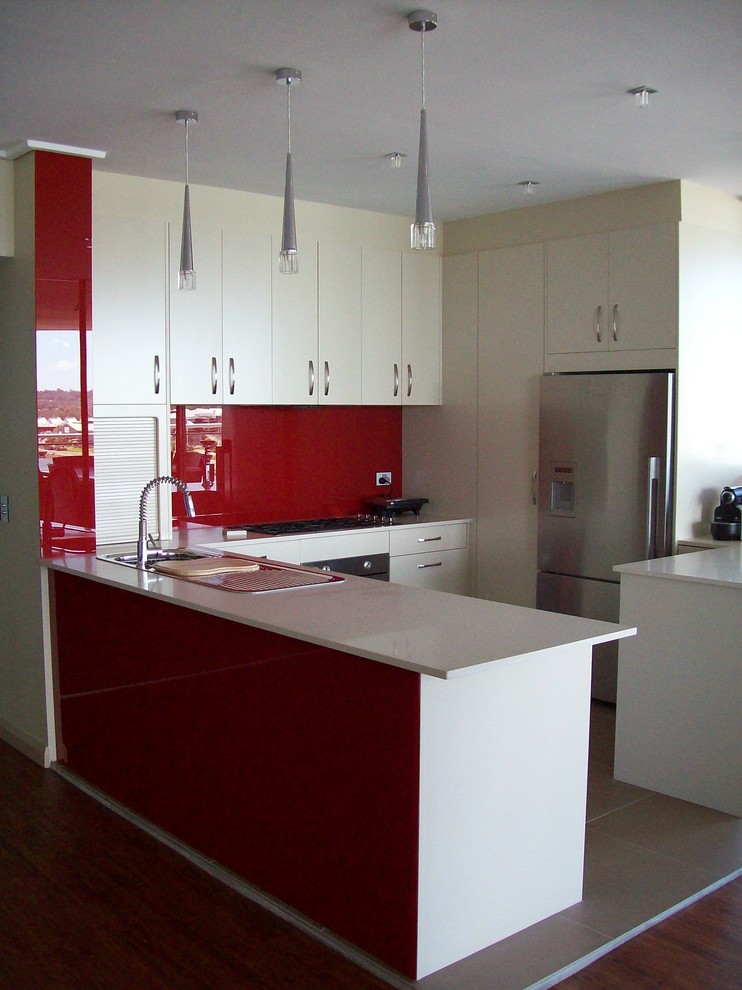 Design ideas for a mid-sized modern u-shaped kitchen in Sydney with a drop-in sink, white cabinets, quartz benchtops, red splashback, glass sheet splashback, stainless steel appliances, porcelain floors and a peninsula.