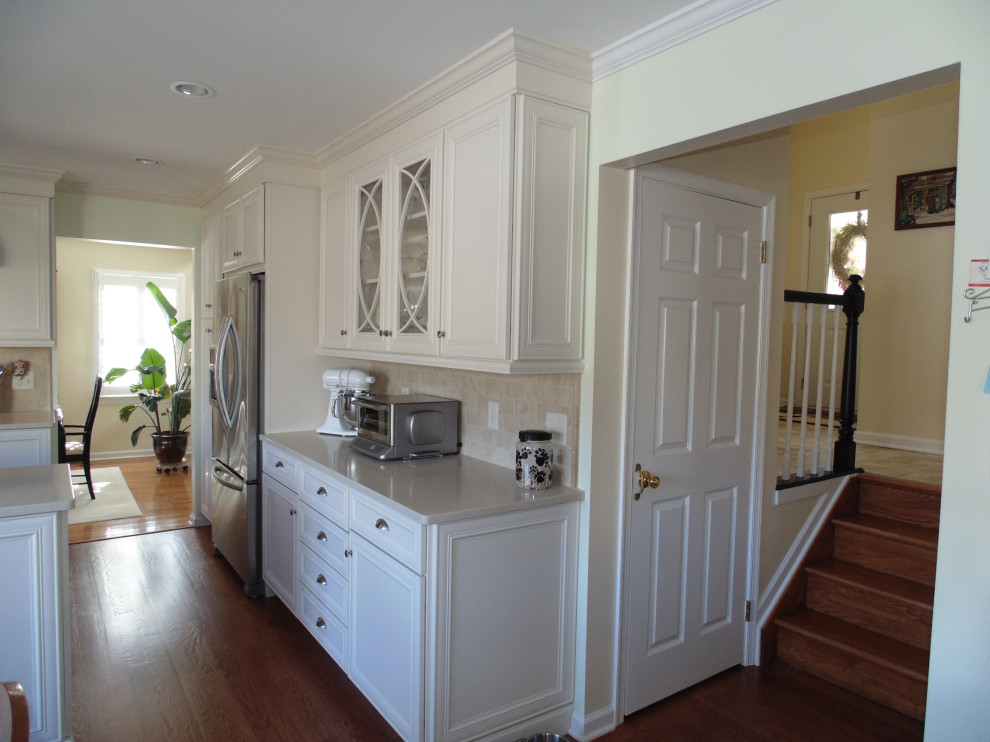 Inspiration for a large transitional u-shaped medium tone wood floor and brown floor eat-in kitchen remodel with a farmhouse sink, flat-panel cabinets, white cabinets, quartz countertops, beige backsplash, travertine backsplash, stainless steel appliances and beige countertops