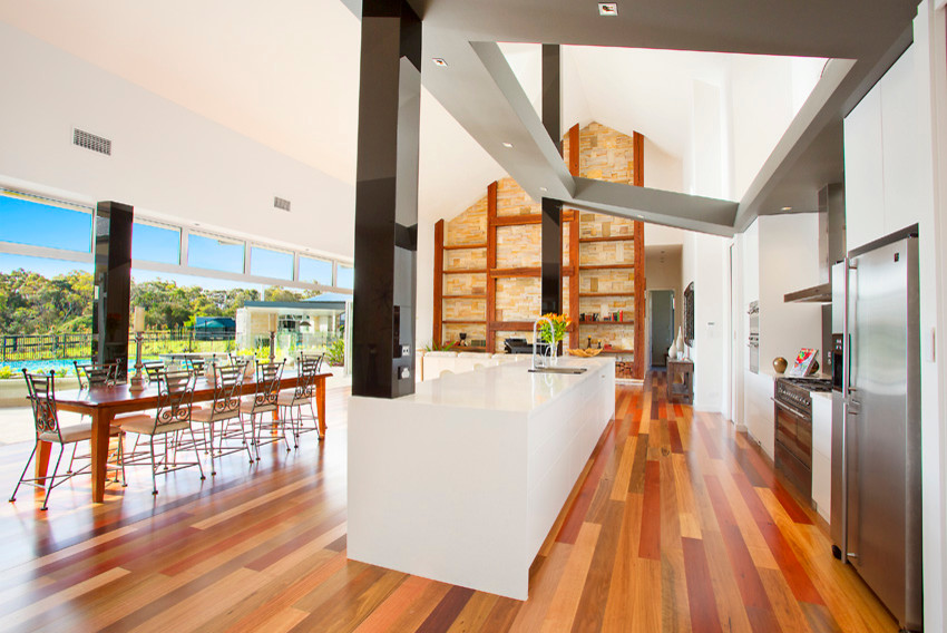 Photo of a large country galley open plan kitchen in Sydney with an undermount sink, stainless steel appliances, medium hardwood floors, with island, flat-panel cabinets, white cabinets, quartz benchtops, black splashback, glass sheet splashback, brown floor and white benchtop.