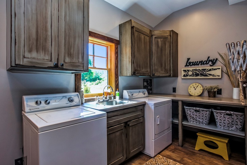 This is an example of a large industrial l-shaped dedicated laundry room in Grand Rapids with a drop-in sink, recessed-panel cabinets, distressed cabinets, laminate benchtops, grey walls, vinyl floors, a side-by-side washer and dryer and brown floor.