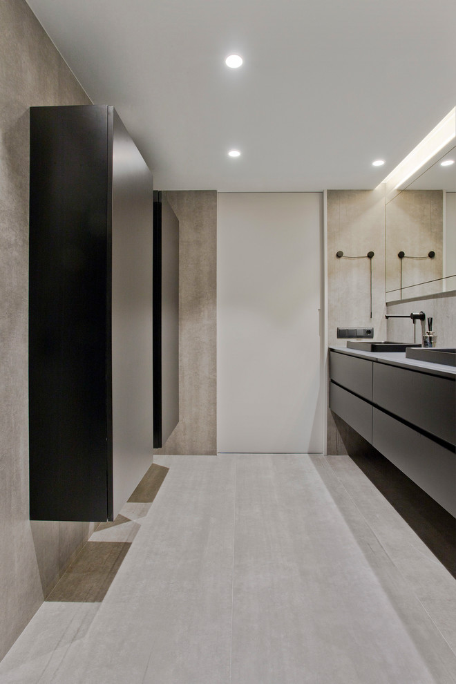 Photo of a mid-sized contemporary powder room in Other with flat-panel cabinets, black cabinets, a wall-mount toilet, gray tile, ceramic tile, grey walls, ceramic floors, a vessel sink, glass benchtops, grey floor and grey benchtops.