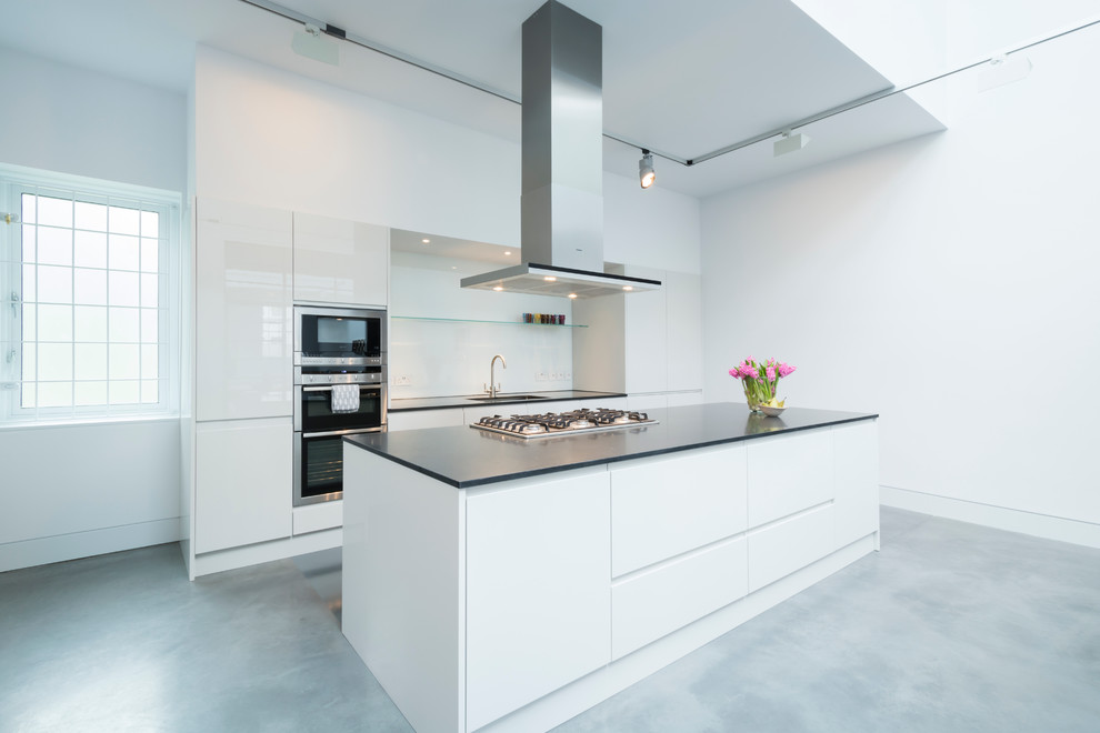 Design ideas for an expansive industrial galley eat-in kitchen in London with with island, a drop-in sink, flat-panel cabinets, solid surface benchtops, white splashback, stainless steel appliances, concrete floors and glass sheet splashback.