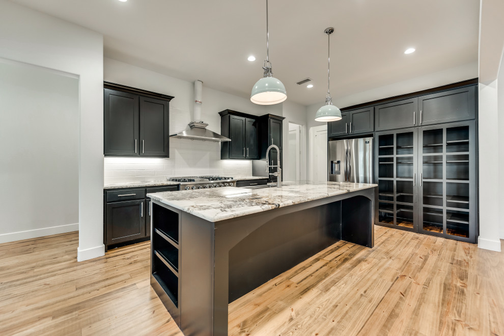 Photo of a mid-sized country l-shaped open plan kitchen in Dallas with glass-front cabinets, black cabinets, granite benchtops, white splashback, subway tile splashback, stainless steel appliances, medium hardwood floors, with island, brown floor, grey benchtop and a farmhouse sink.