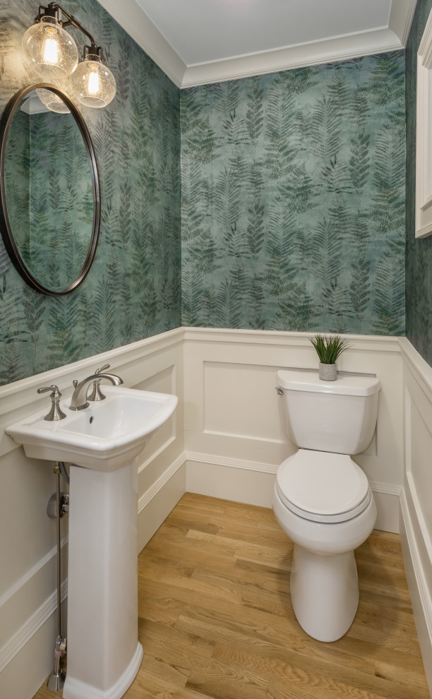 Photo of a small classic cloakroom in Providence with white cabinets, a two-piece toilet, green walls, medium hardwood flooring, a pedestal sink, beige floors and wallpapered walls.