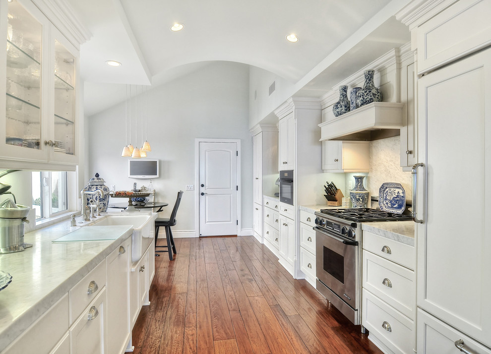 Design ideas for a mid-sized traditional galley separate kitchen in Orange County with a farmhouse sink, recessed-panel cabinets, marble benchtops, white splashback, stone slab splashback, panelled appliances, medium hardwood floors, white cabinets and no island.