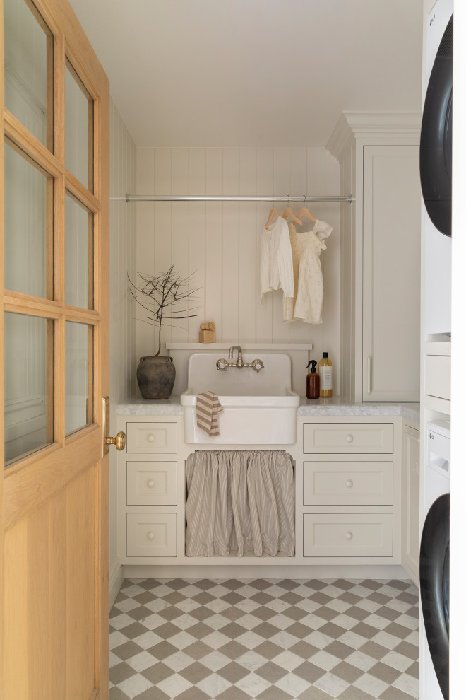 Dedicated laundry room - coastal l-shaped marble floor, multicolored floor and shiplap wall dedicated laundry room idea in Orange County with a farmhouse sink, beige cabinets, marble countertops, shiplap backsplash, beige walls, a stacked washer/dryer, shaker cabinets and gray countertops