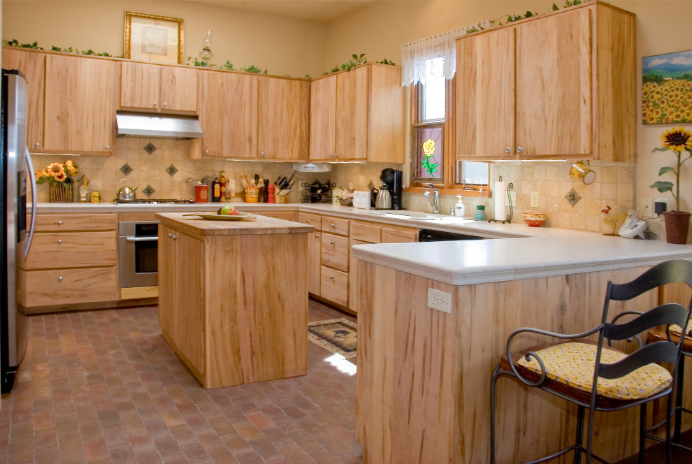 Photo of a large transitional u-shaped eat-in kitchen in Albuquerque with a drop-in sink, flat-panel cabinets, light wood cabinets, tile benchtops, beige splashback, porcelain splashback, stainless steel appliances, brick floors, with island, red floor and white benchtop.