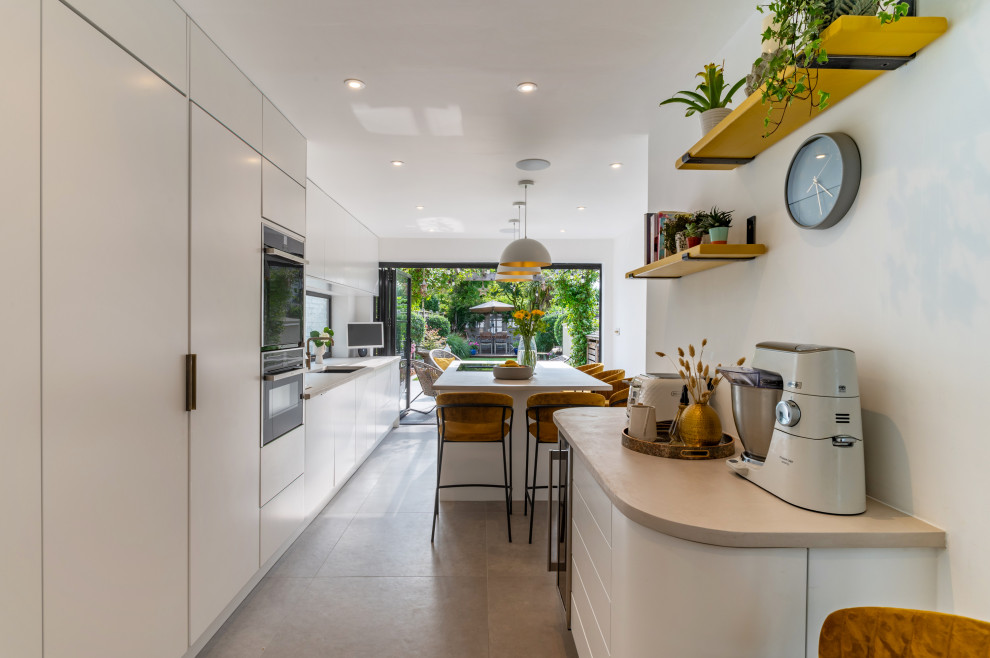 Large contemporary grey and white single-wall open plan kitchen in London with flat-panel cabinets, white cabinets, black appliances, porcelain flooring, multiple islands, grey floors, white worktops and feature lighting.