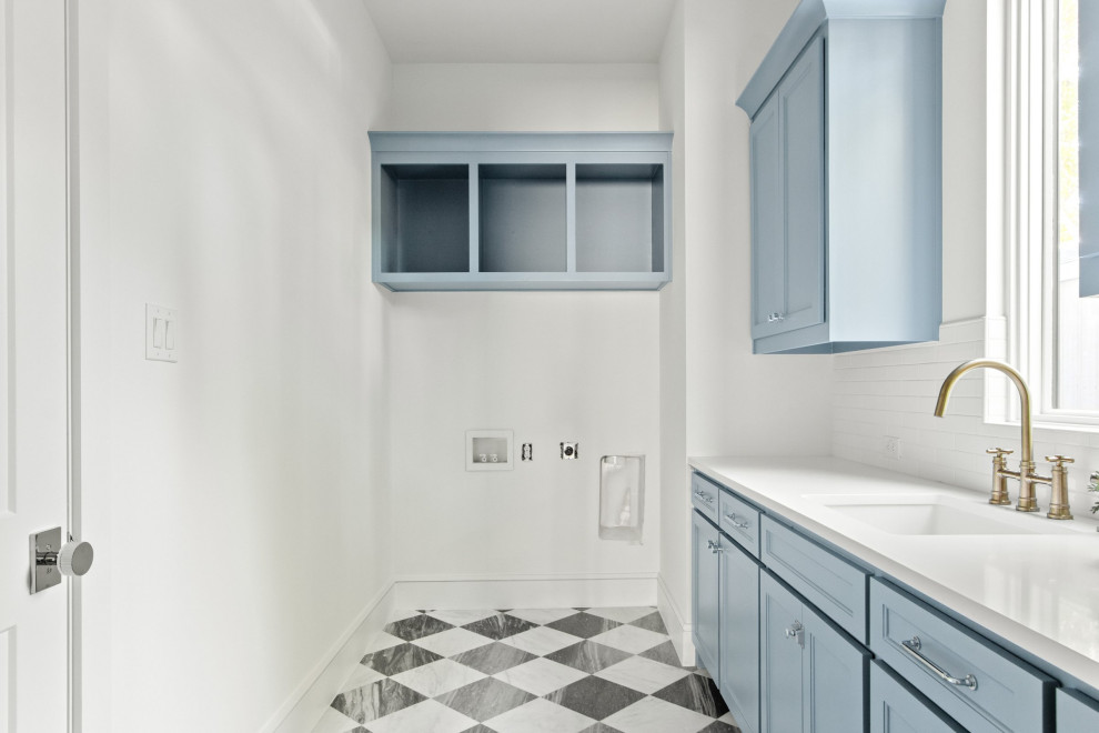 Idéer för maritima linjära vitt tvättstugor enbart för tvätt, med en undermonterad diskho, luckor med profilerade fronter, blå skåp, marmorbänkskiva, vitt stänkskydd, stänkskydd i porslinskakel, vita väggar, marmorgolv, en tvättmaskin och torktumlare bredvid varandra och vitt golv