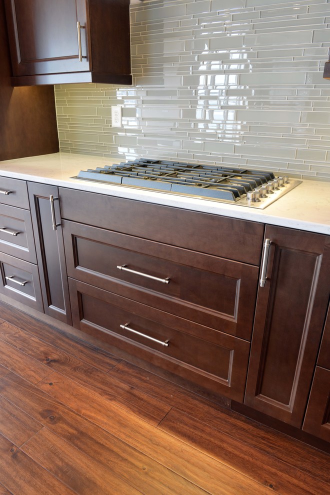 Large transitional l-shaped dark wood floor and brown floor eat-in kitchen photo with an undermount sink, flat-panel cabinets, dark wood cabinets, quartz countertops, gray backsplash, glass sheet backsplash, stainless steel appliances and an island