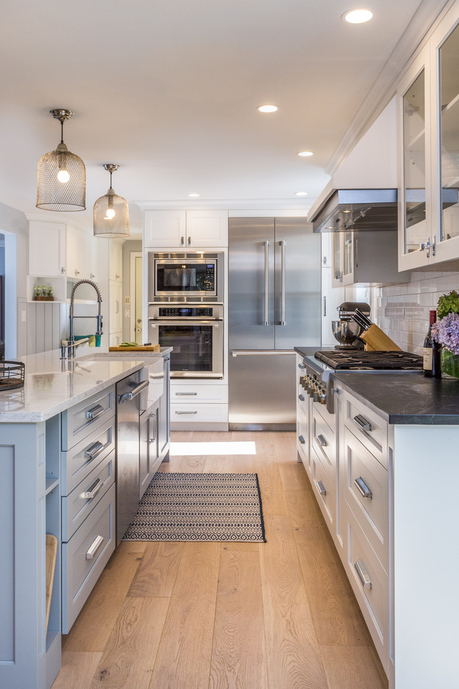 Photo of a transitional kitchen in Manchester with a farmhouse sink, shaker cabinets, white cabinets, white splashback, subway tile splashback, stainless steel appliances, medium hardwood floors, with island and brown floor.