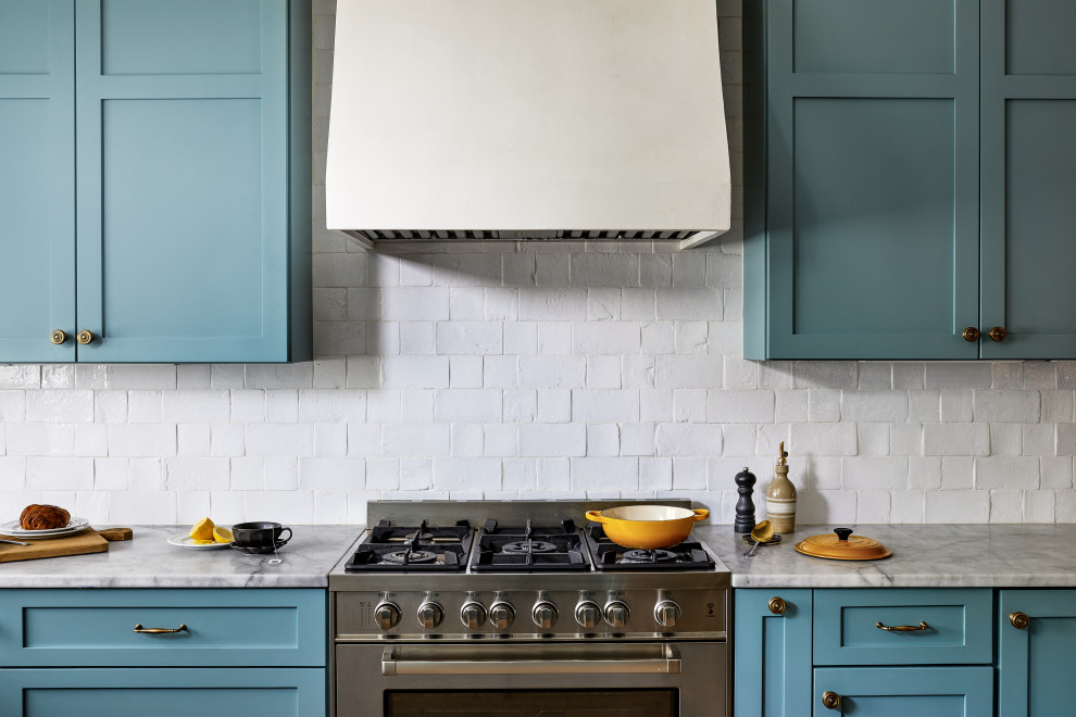 Cette photo montre une grande cuisine américaine chic avec un placard à porte shaker, des portes de placard bleues, plan de travail en marbre et une crédence blanche.