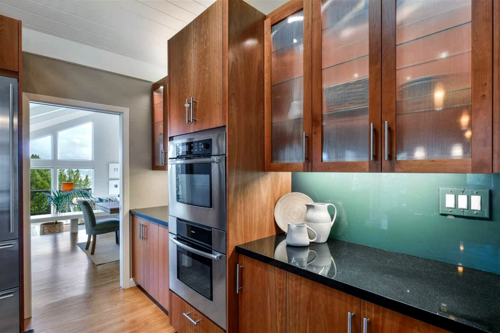 Mid-sized contemporary l-shaped eat-in kitchen in San Francisco with an undermount sink, flat-panel cabinets, dark wood cabinets, granite benchtops, glass sheet splashback, stainless steel appliances, medium hardwood floors, with island, brown floor, black benchtop, exposed beam, vaulted and wood.