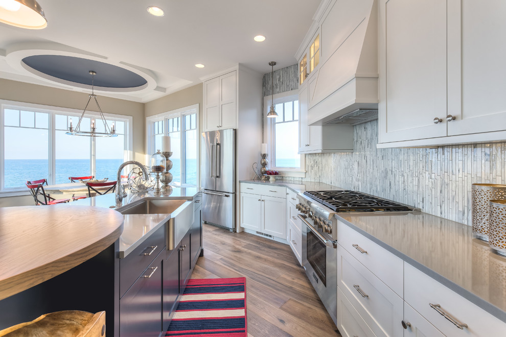 Photo of a mid-sized beach style galley kitchen in Chicago with a farmhouse sink, shaker cabinets, white cabinets, quartzite benchtops, grey splashback, matchstick tile splashback, stainless steel appliances, light hardwood floors, with island and multi-coloured floor.