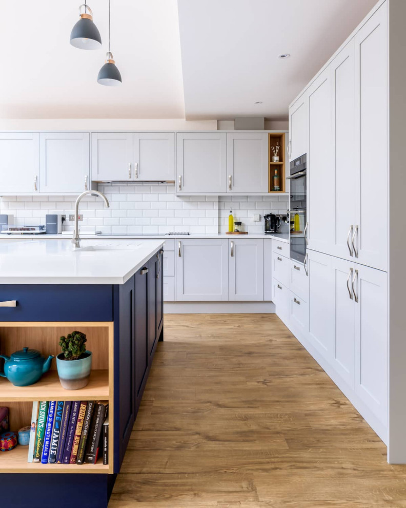 Photo of a large classic l-shaped open plan kitchen in Sussex with a single-bowl sink, shaker cabinets, white cabinets, quartz worktops, white splashback, ceramic splashback, black appliances, light hardwood flooring, an island, brown floors, white worktops, a drop ceiling and feature lighting.