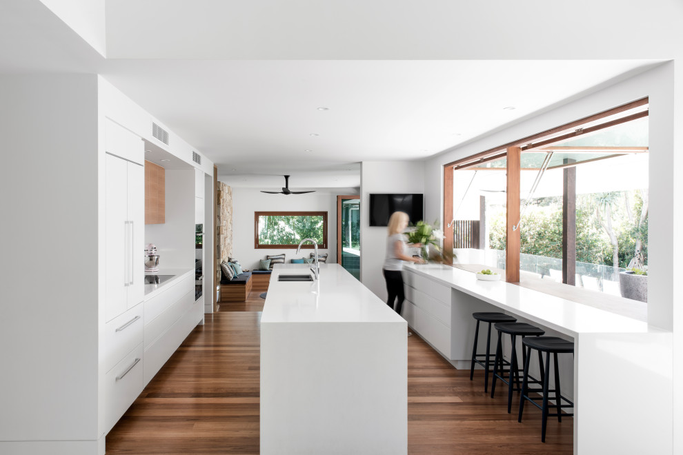 Design ideas for a contemporary galley kitchen in Sunshine Coast with an undermount sink, flat-panel cabinets, white cabinets, panelled appliances, medium hardwood floors, with island, brown floor and white benchtop.
