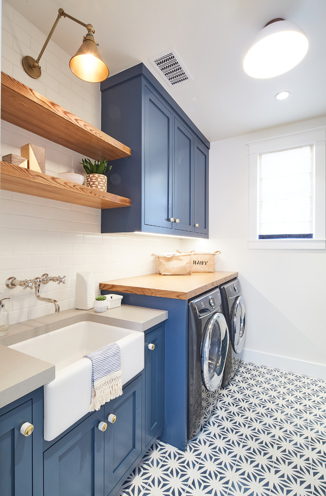 Photo of a beach style single-wall dedicated laundry room in Los Angeles with a farmhouse sink, shaker cabinets, blue cabinets, wood benchtops, white walls, a side-by-side washer and dryer, multi-coloured floor and grey benchtop.