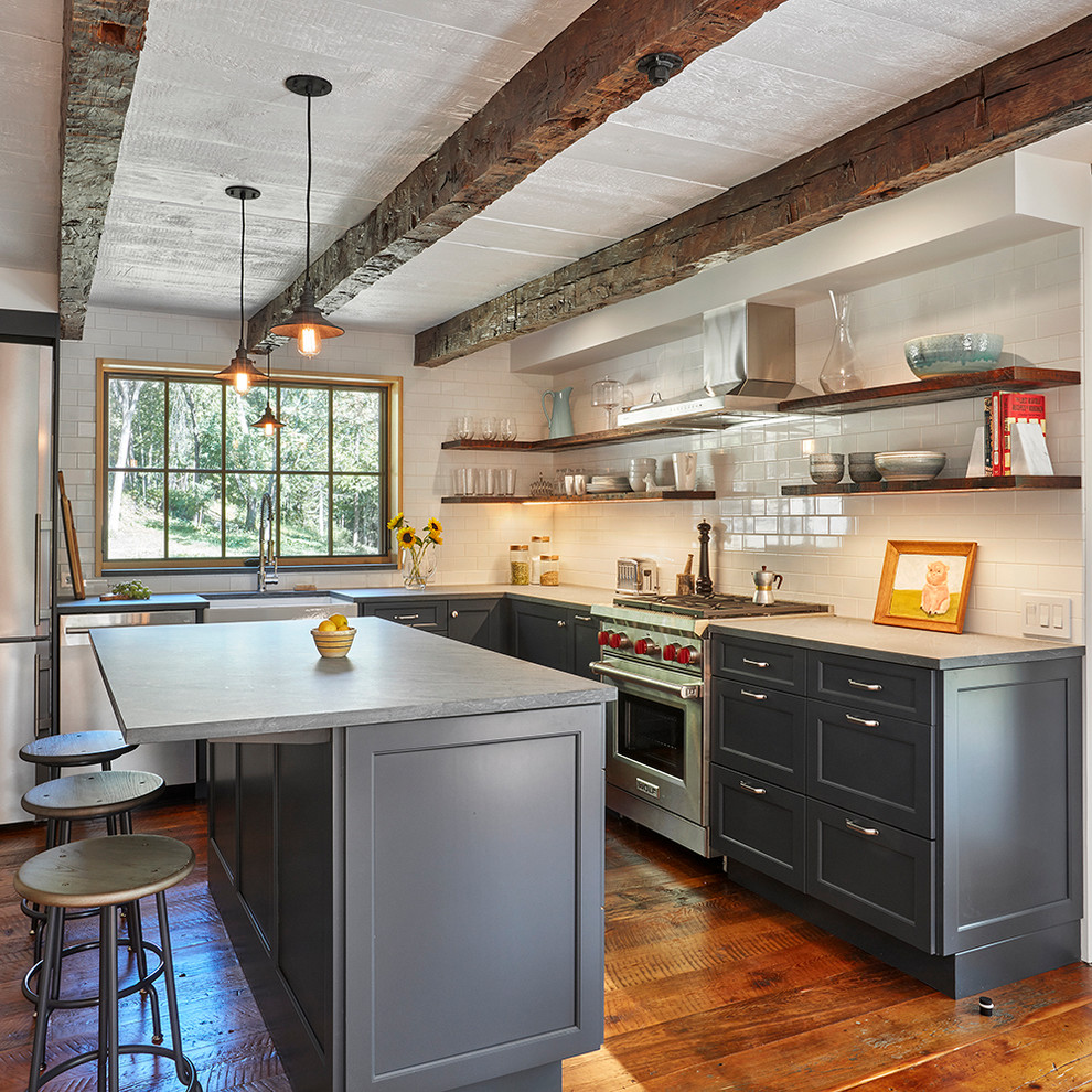 This is an example of a country l-shaped kitchen in New York with a farmhouse sink, shaker cabinets, grey cabinets, white splashback, subway tile splashback, stainless steel appliances, medium hardwood floors, with island and brown floor.