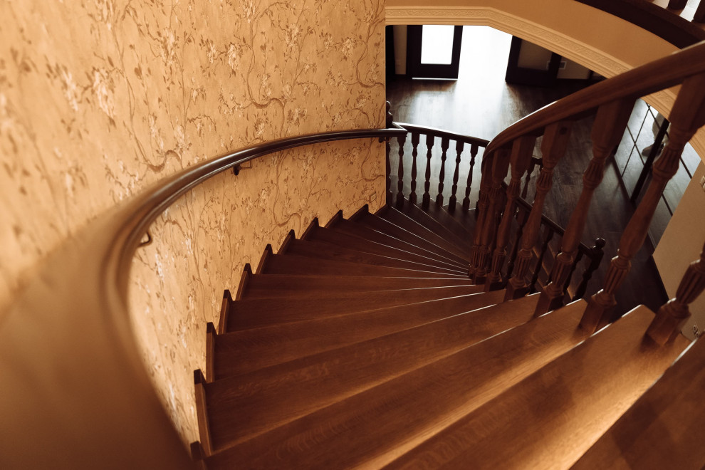 На фото: большая изогнутая деревянная лестница в классическом стиле с деревянными ступенями, деревянными перилами, обоями на стенах и кладовкой или шкафом под ней с