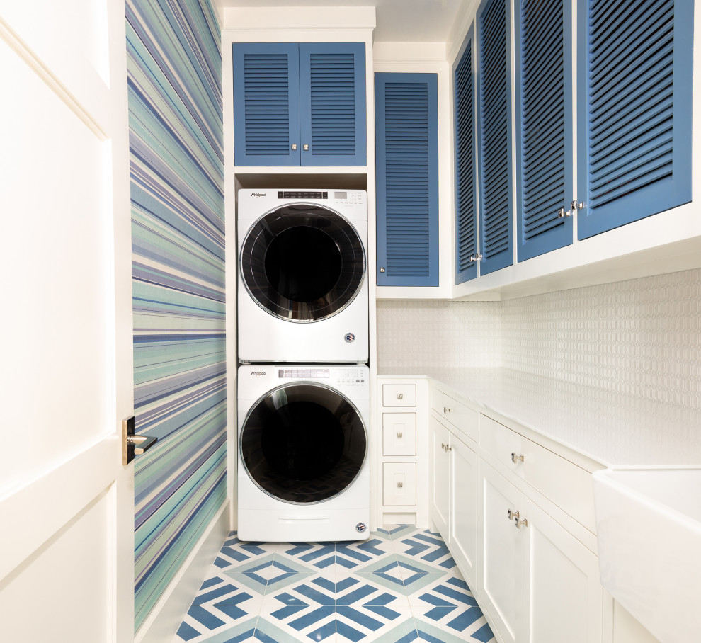 Design ideas for a transitional l-shaped laundry room in Dallas with a farmhouse sink, louvered cabinets, blue cabinets, multi-coloured walls, a stacked washer and dryer, multi-coloured floor, white benchtop and wallpaper.