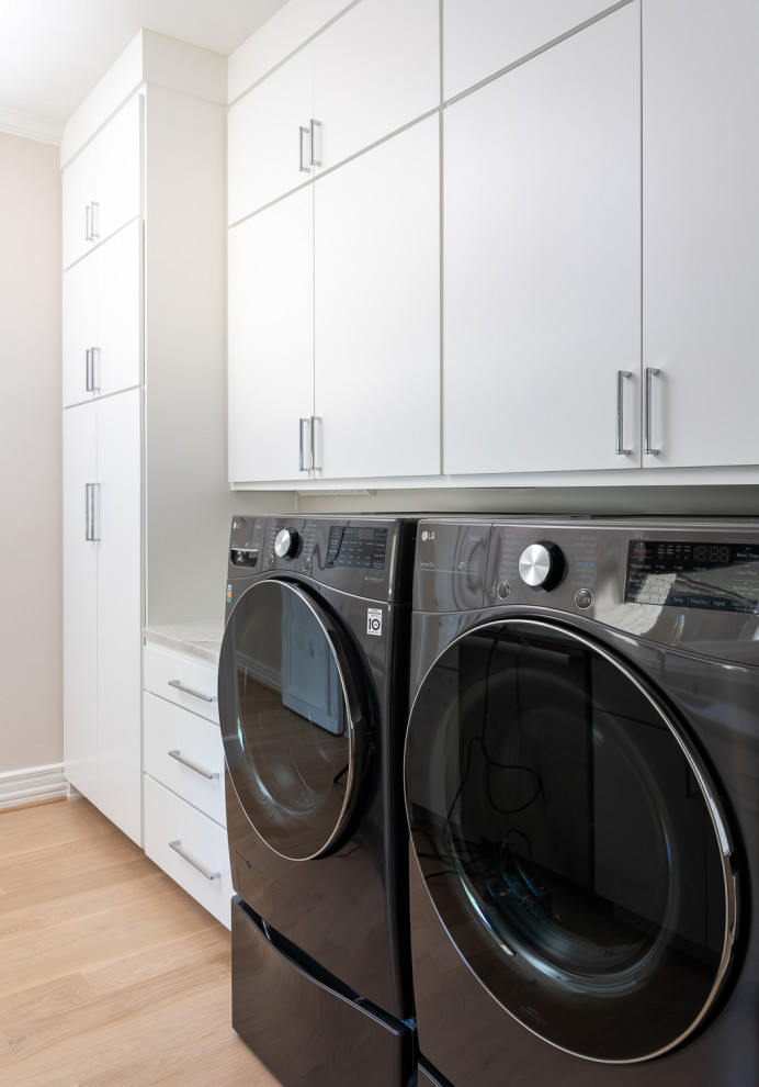 Modern inredning av en vita parallell vitt tvättstuga, med en undermonterad diskho, släta luckor, vita skåp, bänkskiva i kvartsit, grå väggar, ljust trägolv och en tvättmaskin och torktumlare bredvid varandra