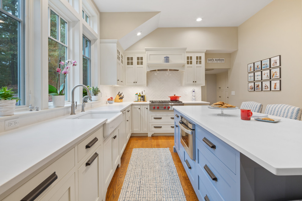 Inspiration for a large coastal l-shaped medium tone wood floor and brown floor kitchen pantry remodel in Boston with a farmhouse sink, shaker cabinets, white cabinets, quartz countertops, white backsplash, ceramic backsplash, stainless steel appliances, an island and white countertops