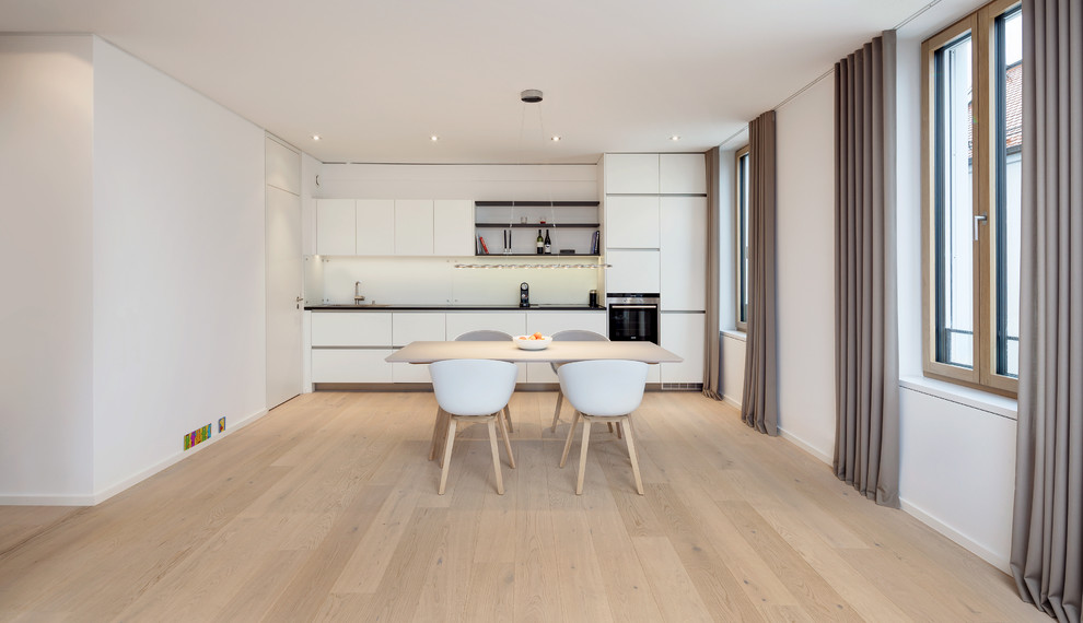 This is an example of a mid-sized modern single-wall open plan kitchen in Munich with a single-bowl sink, flat-panel cabinets, white cabinets, white splashback, black appliances, light hardwood floors and no island.