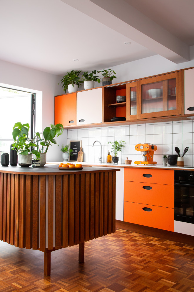 Mittelgroße Retro Küche mit flächenbündigen Schrankfronten, Mineralwerkstoff-Arbeitsplatte, Küchenrückwand in Weiß, Rückwand aus Keramikfliesen, dunklem Holzboden, Kücheninsel, weißer Arbeitsplatte, orangefarbenen Schränken und freigelegten Dachbalken in Sussex