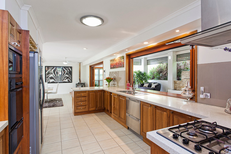 Photo of a mid-sized traditional galley open plan kitchen in Brisbane with a double-bowl sink, recessed-panel cabinets, medium wood cabinets, granite benchtops, grey splashback, glass tile splashback, stainless steel appliances, travertine floors and a peninsula.