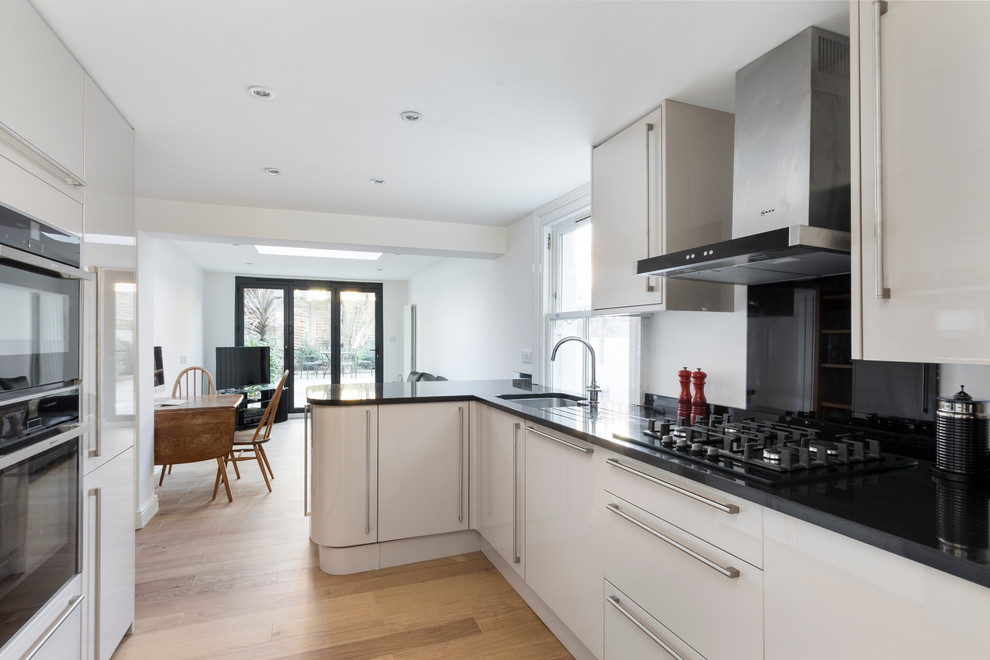 Photo of a mid-sized contemporary l-shaped open plan kitchen in London with a drop-in sink, shaker cabinets, white cabinets, granite benchtops, black splashback, stainless steel appliances and light hardwood floors.