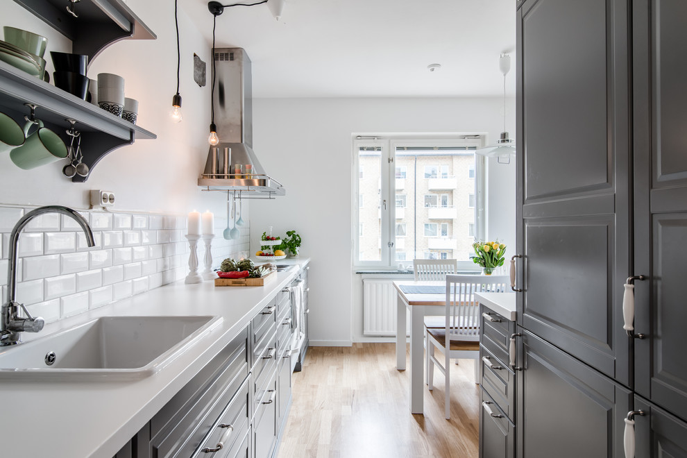 This is an example of a mid-sized scandinavian galley eat-in kitchen in Stockholm with a drop-in sink, raised-panel cabinets, grey cabinets, white splashback, subway tile splashback, light hardwood floors and no island.