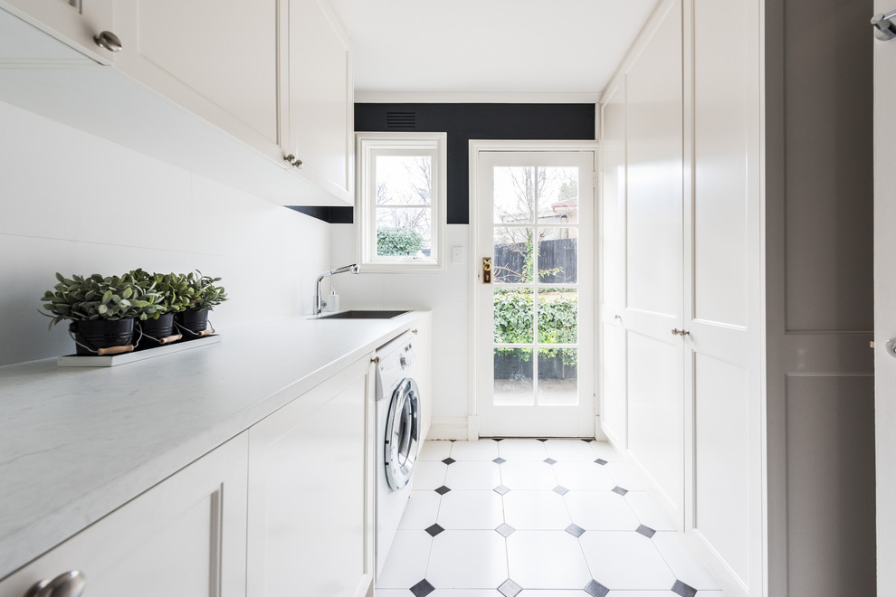 This is an example of a mid-sized contemporary single-wall dedicated laundry room in Melbourne with shaker cabinets, white cabinets, quartz benchtops, black walls, ceramic floors, a side-by-side washer and dryer, multi-coloured floor, white benchtop and an undermount sink.