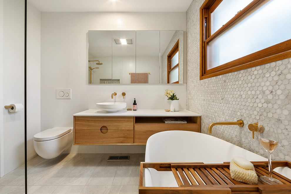 Photo of a mid-sized contemporary kids bathroom in Melbourne with furniture-like cabinets, medium wood cabinets, a freestanding tub, an open shower, a wall-mount toilet, gray tile, mosaic tile, white walls, porcelain floors, a vessel sink, engineered quartz benchtops, grey floor, an open shower and white benchtops.