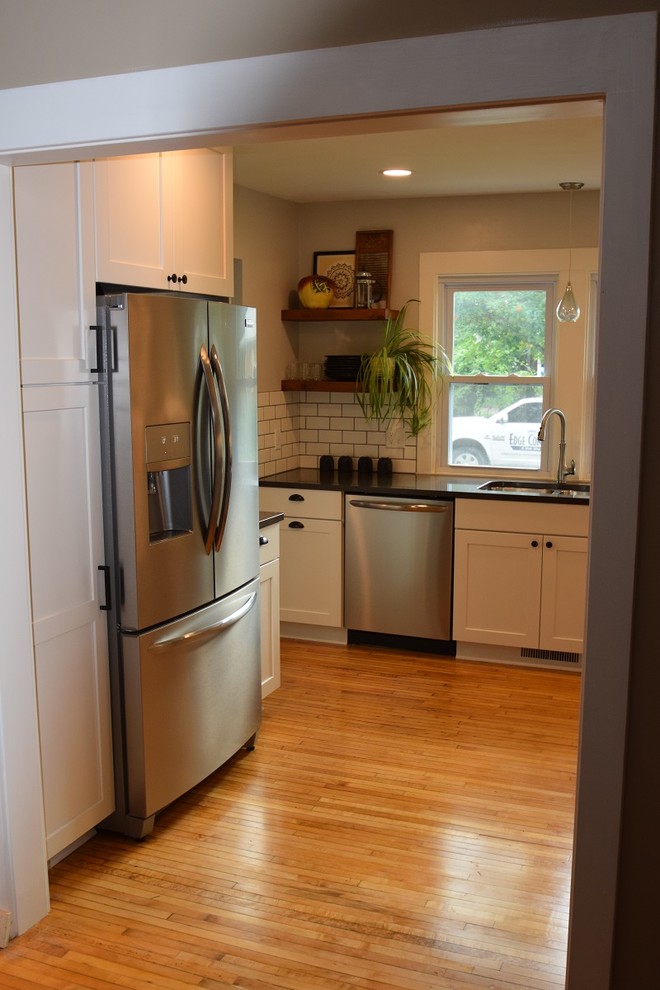 Example of a transitional u-shaped light wood floor kitchen design with an undermount sink, shaker cabinets, white cabinets, quartz countertops, white backsplash, subway tile backsplash, stainless steel appliances, no island and black countertops