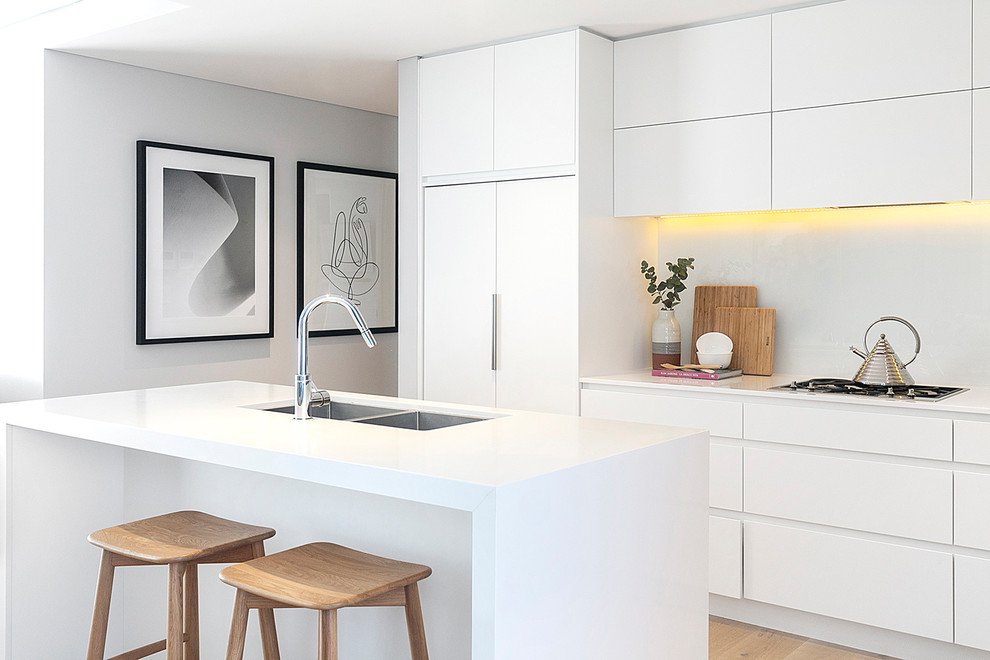 This is an example of a contemporary galley kitchen in Sydney with a double-bowl sink, flat-panel cabinets, white cabinets, white splashback, panelled appliances, with island, white benchtop, light hardwood floors and beige floor.
