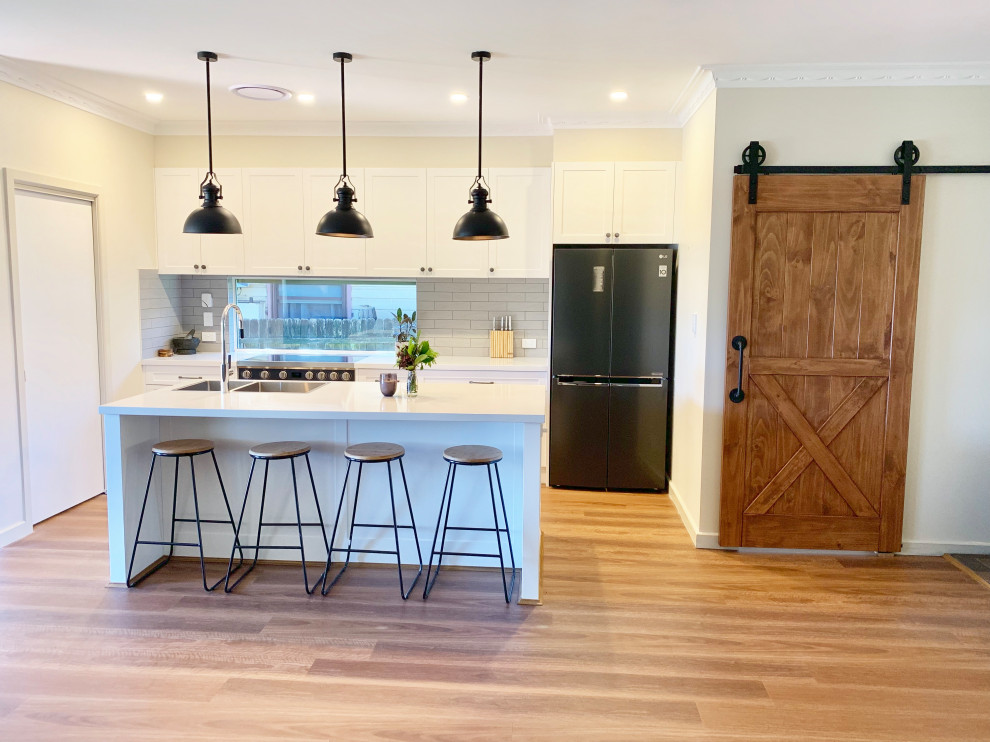 This is an example of a mid-sized contemporary galley open plan kitchen in Sydney with a double-bowl sink, shaker cabinets, white cabinets, quartz benchtops, grey splashback, subway tile splashback, black appliances, light hardwood floors, with island, brown floor and white benchtop.